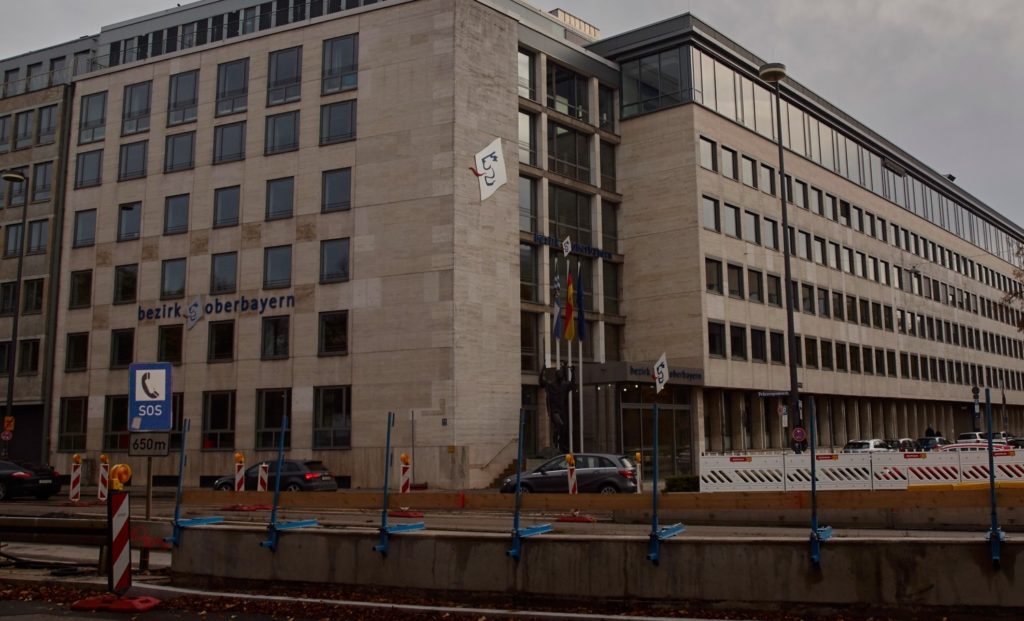 Das Bild zeigt das Gebäude der Behörde "Bezirk Oberbayern". Im Vordergrund ein SOS-Schild.